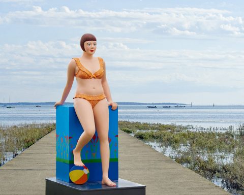 Femme en maillot de bain orange sur un socle coloré au bord de l'eau.